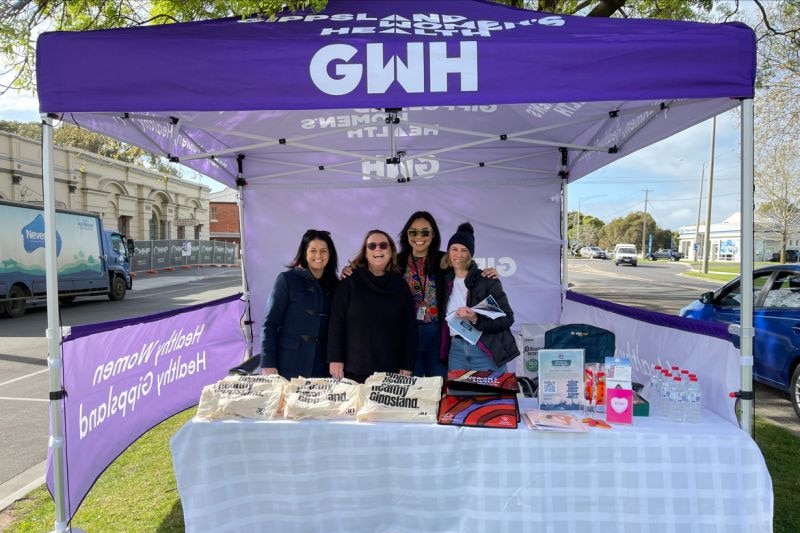 Four women stand underneath a gazebo tent with a logo that reads Gippsland Women's Health Stall 