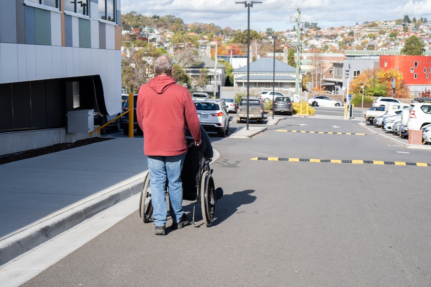 A man in a wheelchair is photographed with his back to the camera, while a tall man in a red hoodie wheels him down a road.