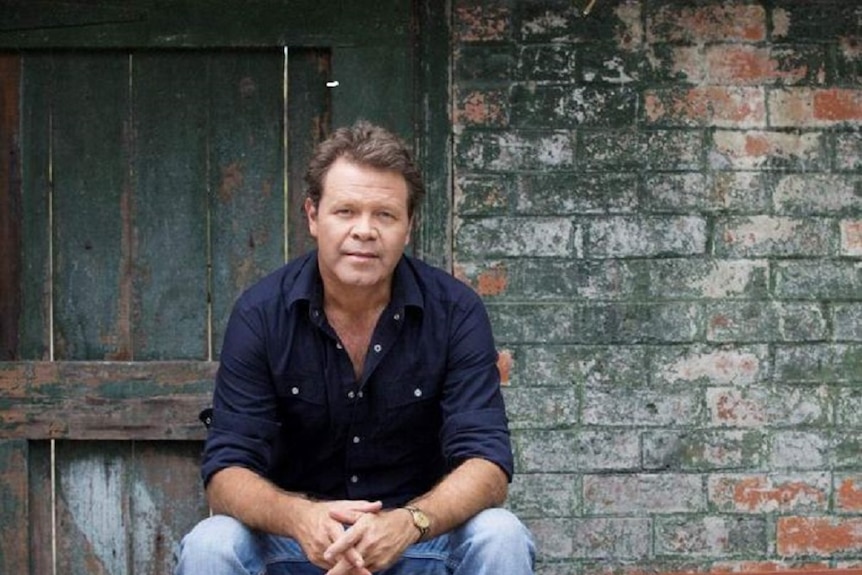 Musician Troy Cassar-Daley sits on a wooden bench, a solemn look on his face.