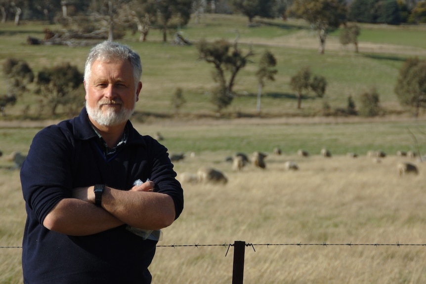 A grey-bearded man in a deep navy blue jumper, with arms crossed, standing in front of a barbed wire fence, sheep in distance