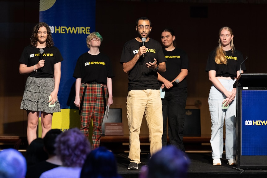 Five young people on stage in black 'heywire' shirts. A young man in front with a microphone.