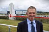 Grey haired man in navy suit smiling in front of Bong Bong Picnic Races sign and track