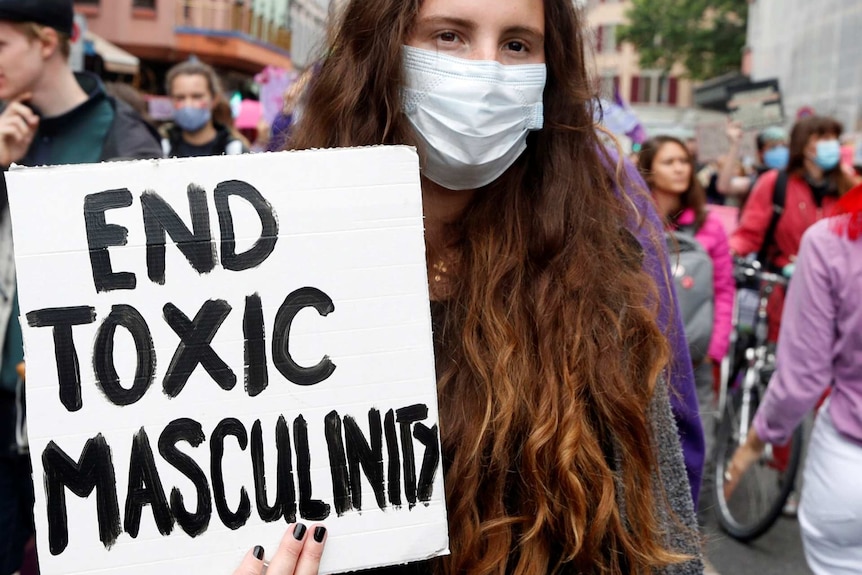 a woman with long red hair wearing a mask holds up a sign saying end toxic masculinity