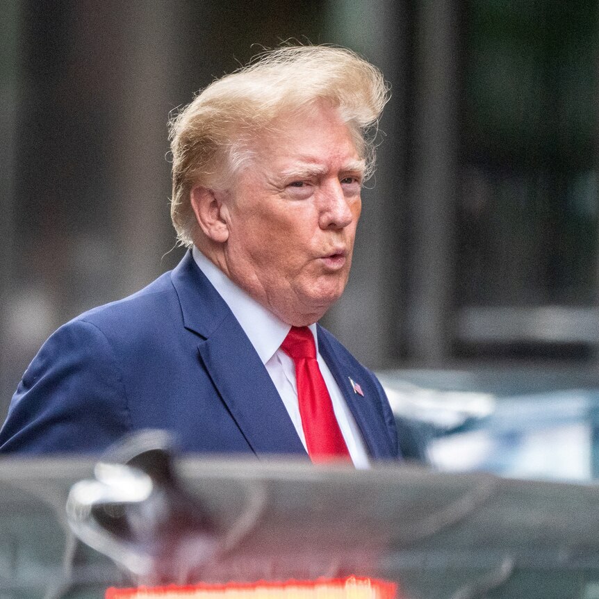 Donald Trump wearing a blue suit and red tie walks to a car with his hair blowing in the wind.