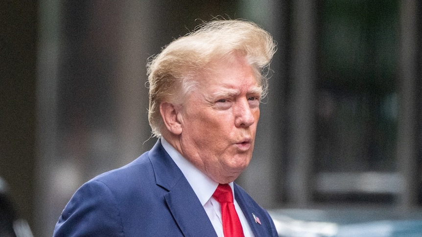 Donald Trump wearing a blue suit and red tie walks to a car with his hair blowing in the wind.