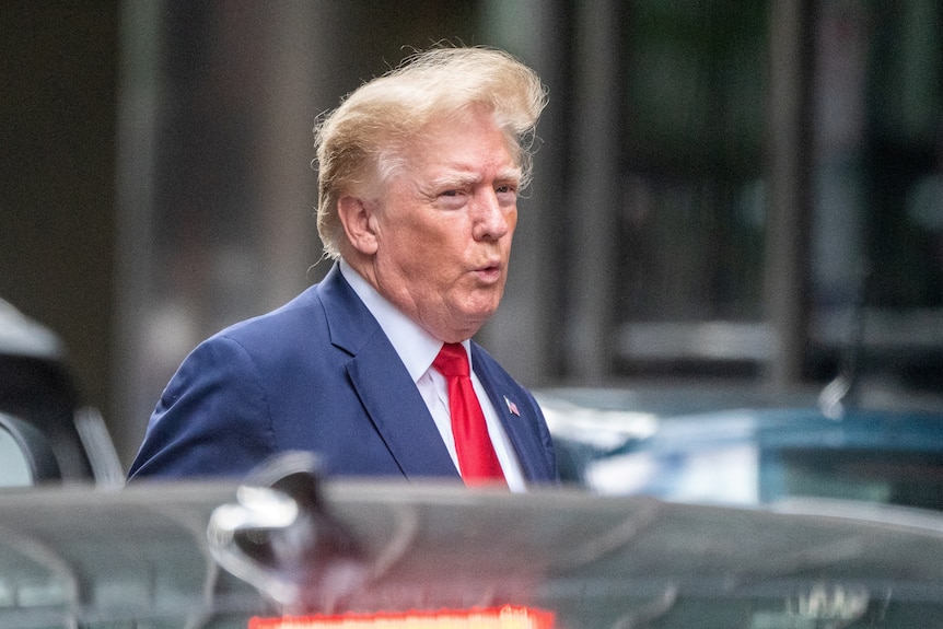 Donald Trump wearing a blue suit and red tie walks to a car with his hair blowing in the wind.