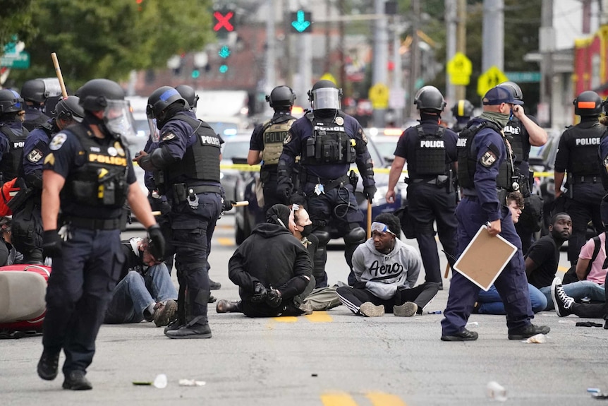Police detain a group of people as they sit on the ground with their hands cuffed behind their backs.
