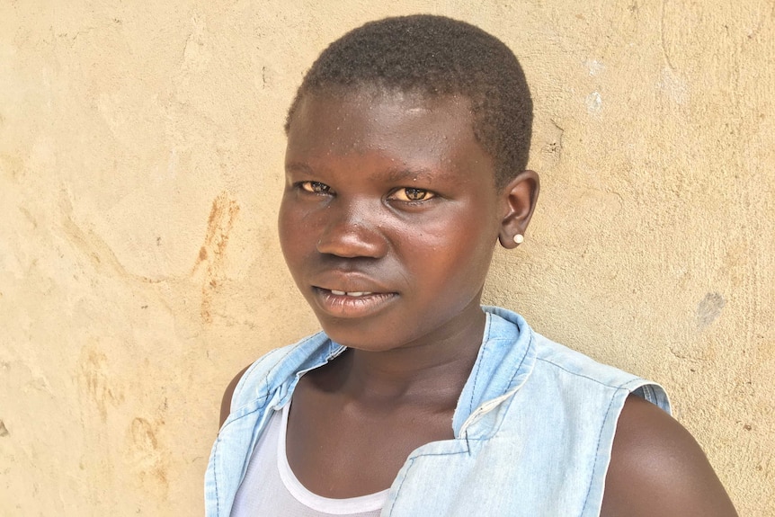 A teenage girl in South Sudan, stands against a brick wall looking into the camera.