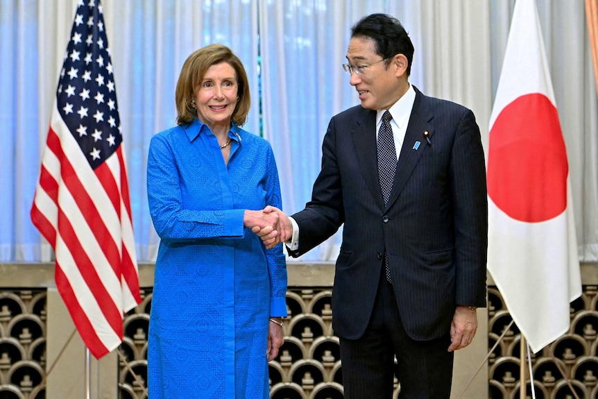 Woman in blue suit shakes hands with man in black suit with US and Japanese flags in background.