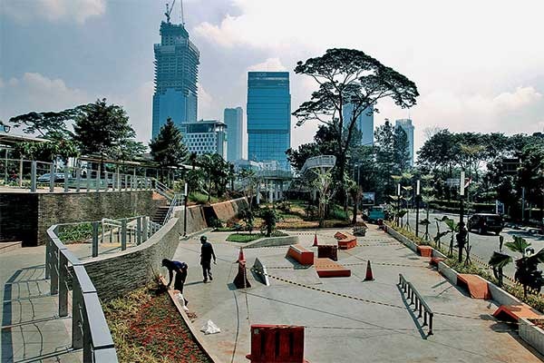A long shot of an open public space with a skate park and a park, in front of skyscrapers.