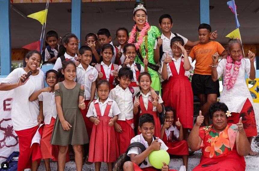 Miss Samoa dressed in green puletasi wears crown, surrounded by young children in white and red school uniform. 