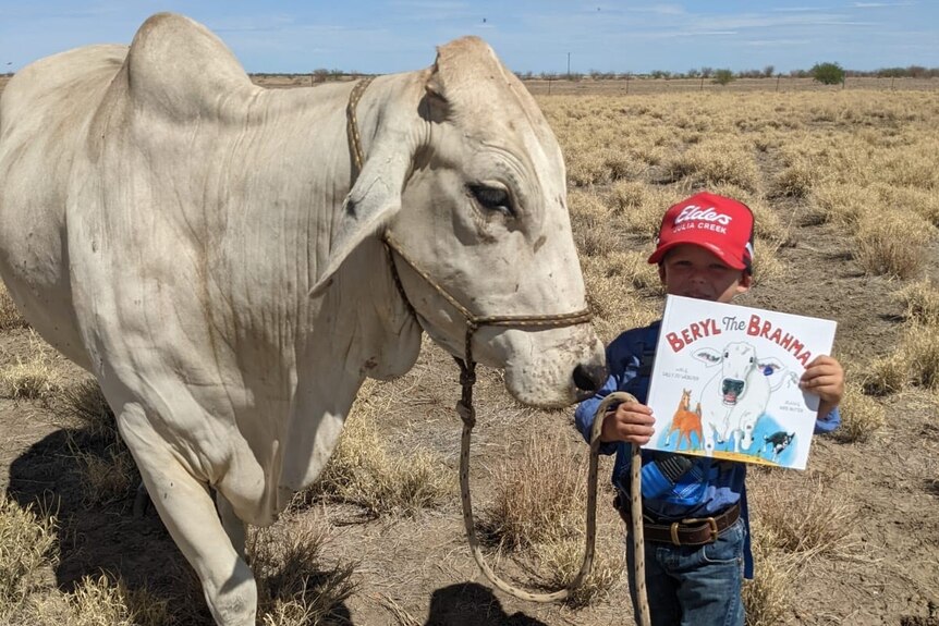 A small boy stands next to a cow holding a kids picture book