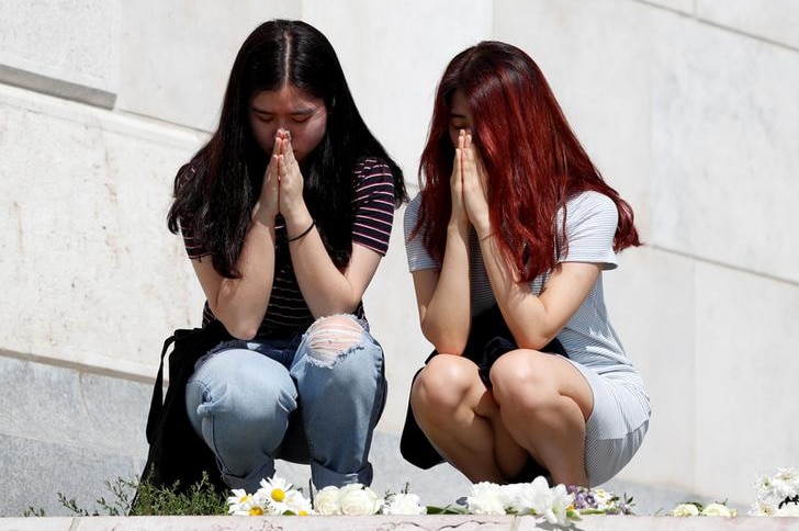 Two women kneel and hold their hands in prayer position to their faces. They are among floral tributes.