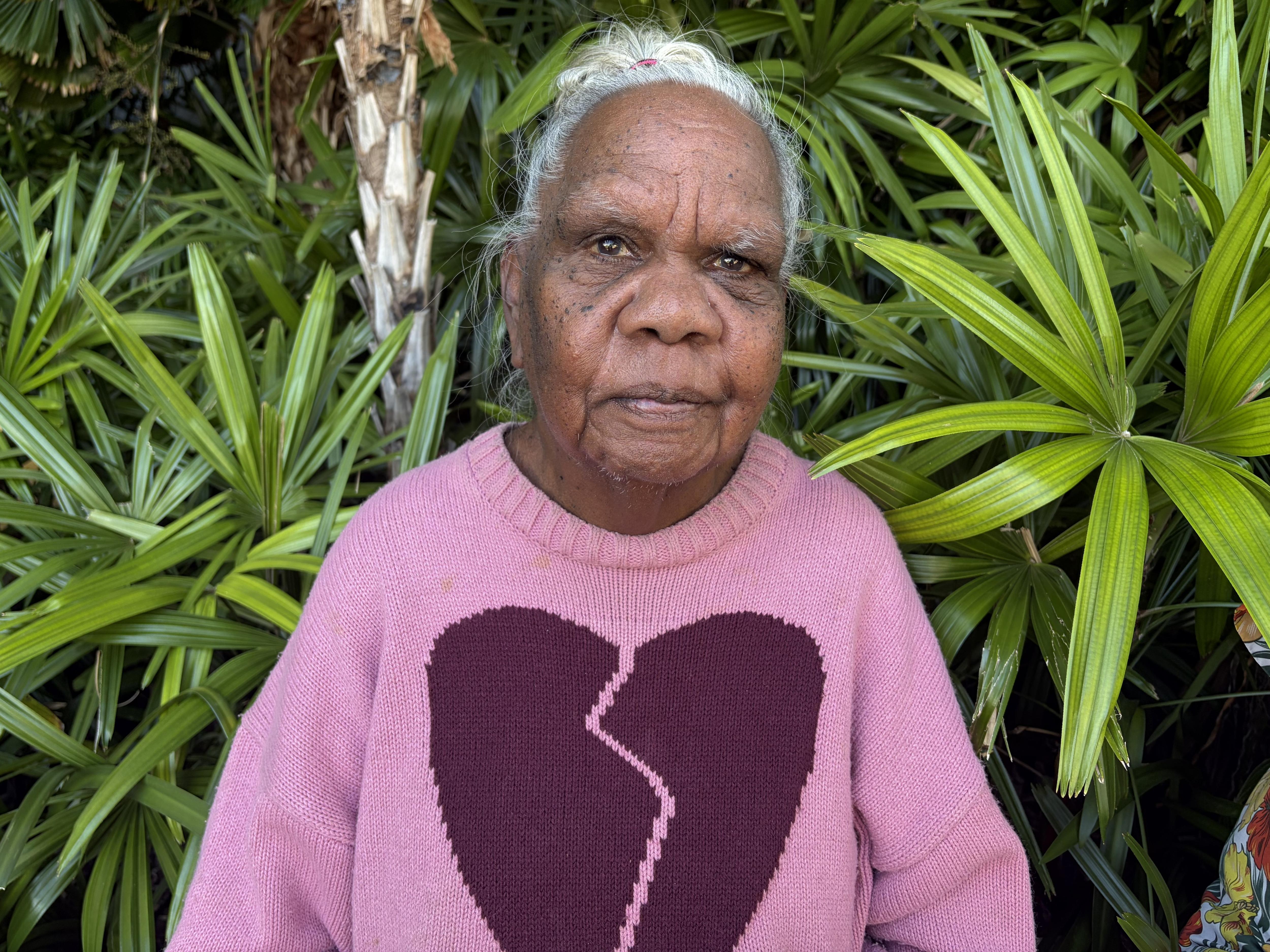 An Aboriginal woman, gray hair, wearing pink jumper, with a broken dark purple heart on the front, standing among green plants