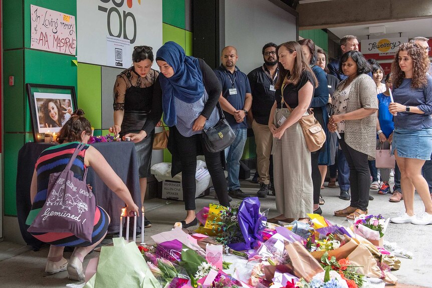 People line up to light candles at a table with a framed photo of Aiia Maasarwe on it, with flowers on the ground nearby.