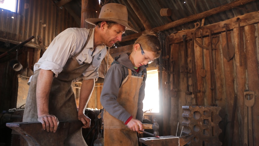 A father looks over his son's shoulder as he uses blacksmithing equipment.