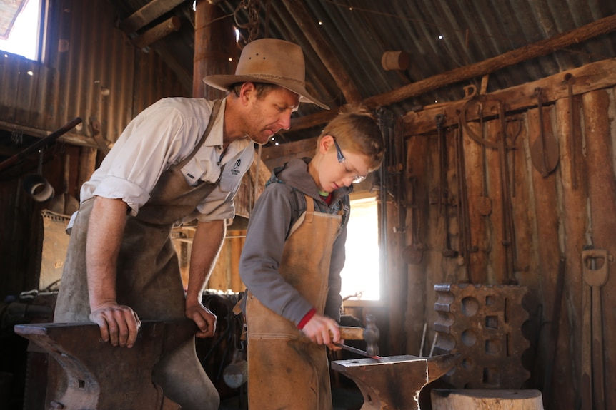 A father looks over his son's shoulder as he uses blacksmithing equipment.