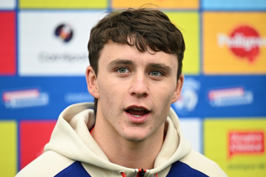A young man with brown hair speaks to the media in front of a sponsorship sign