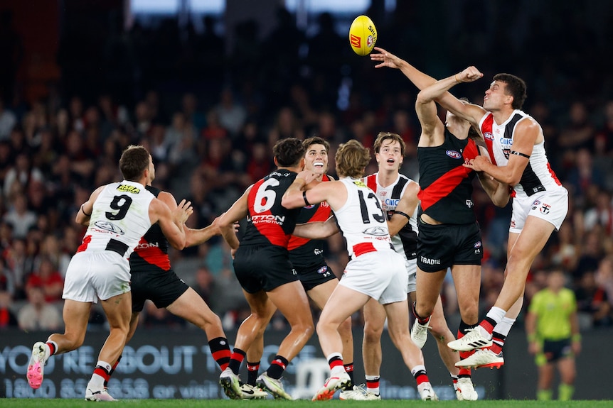 A St Kilda ruckman reaches out to palm the ball away against an Essendon ruckman as other players watch from the ground.
