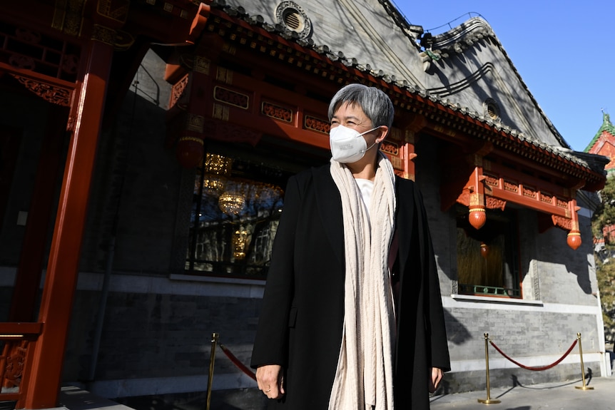 Penny Wong in face mask, coat and scarf stands in front of an ornate Chinese building. 
