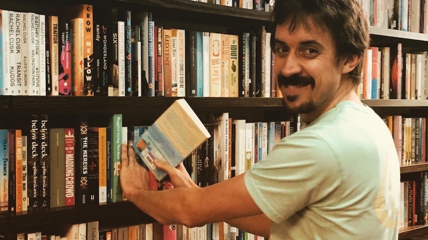 man with moustache wearing a t-shirt placing book in a full book shelf