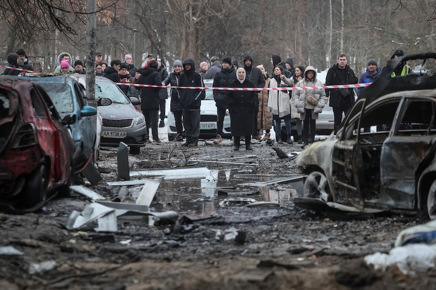 Two dozen residents stand behind a rope, away from debris and burned out cars after a missile strike