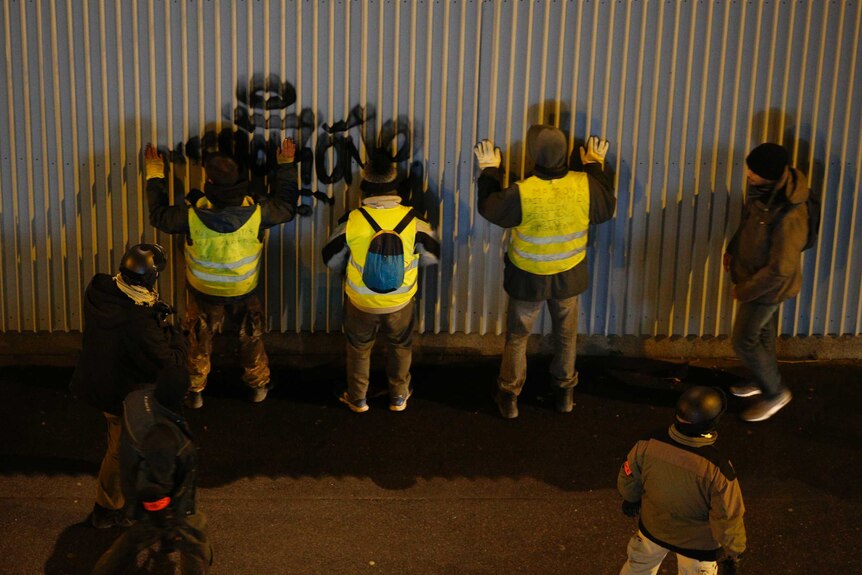 Three yellow vest protesters have their hands up against a corrugated metal wall as police officers search them.
