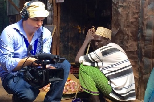 Cuddihy squats down while holding a camera, next to a man sitting on a stool outside a shop front.