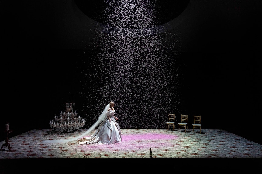 White woman with blonde hair elegantly styled wears white wedding dress and crossed softly lit stage with pink confetti falling