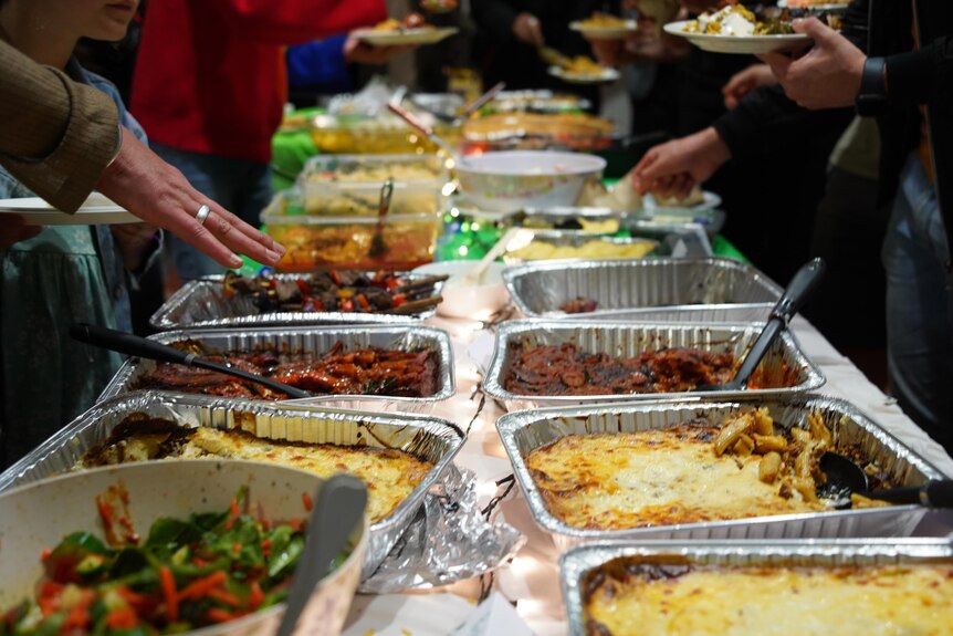 Dozens of metal trays of various foods lined up across a long table, and hands reaching in to serve their plates.