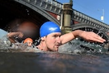 A man in a blue bathing cap pulls his arm out of the river in a free-style motion with the Princes Bridge in the background.