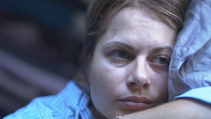 A woman rest her head on a pillow, she looks to the side, with a neutral expression.