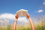 Arms holding a book against a blue sky, surrounded by field of high grass.