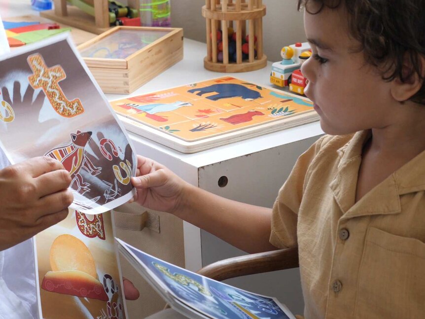 A child wearing a burnt yellow shirt looks at pictures with Indigenous drawings on them in what appears to be a classroom.