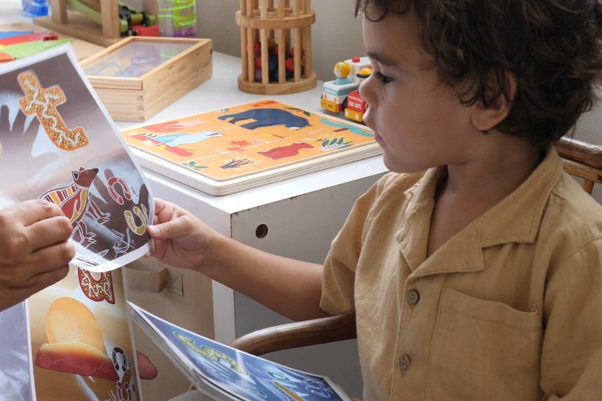 A child wearing a burnt yellow shirt looks at pictures with Indigenous drawings on them in what appears to be a classroom.