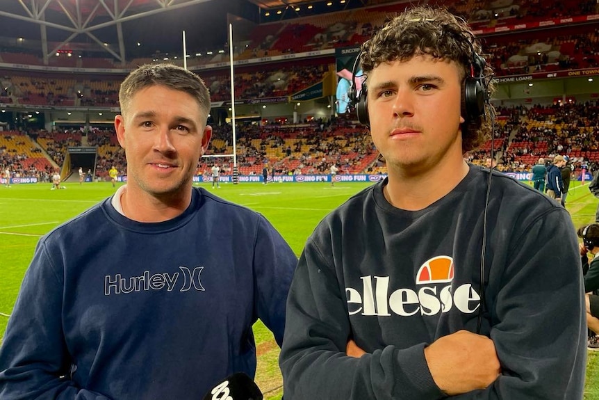 Two young men stand beside each other in dark jumpers, holding a microphone, with the Suncorp stadium football field behind them