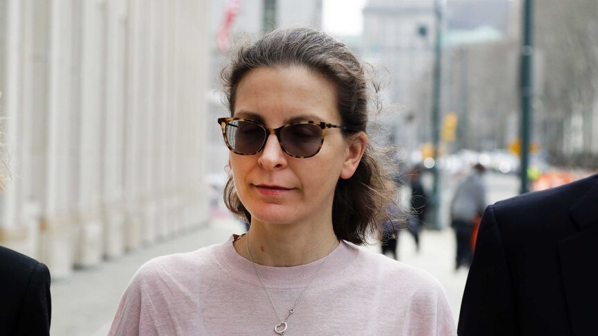 A woman is pictured close-up in a pale pink jumper with a heart-shape item dangling on a necklace walking on an overcast street.