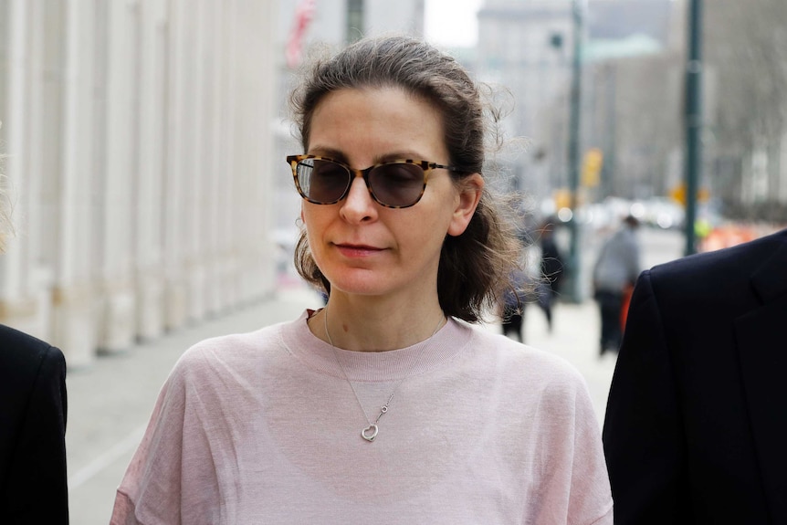 A woman is pictured close-up in a pale pink jumper with a heart-shape item dangling on a necklace walking on an overcast street.