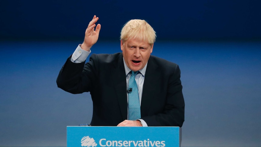 Britain's Prime Minister Boris Johnson holds his hand to the arm as he speaks from a podium.