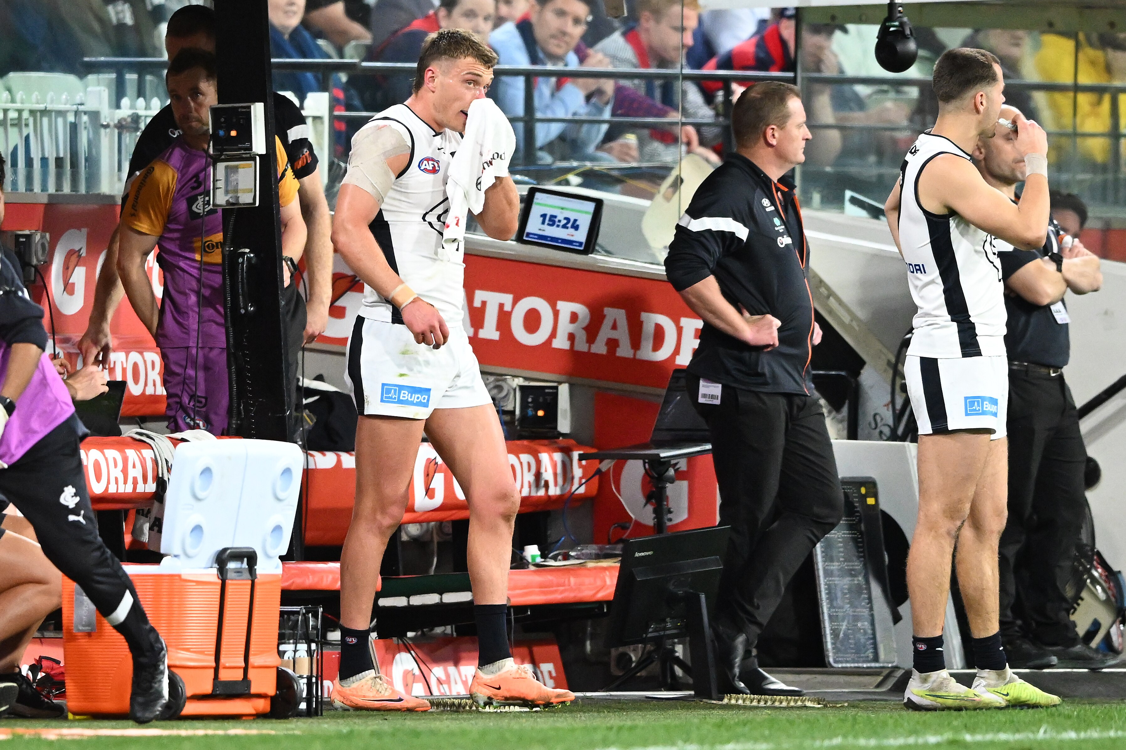Carlton AFL star Patrick Cripps stands near the team dugout during a game, using a towel to wipe his bleeding nose.