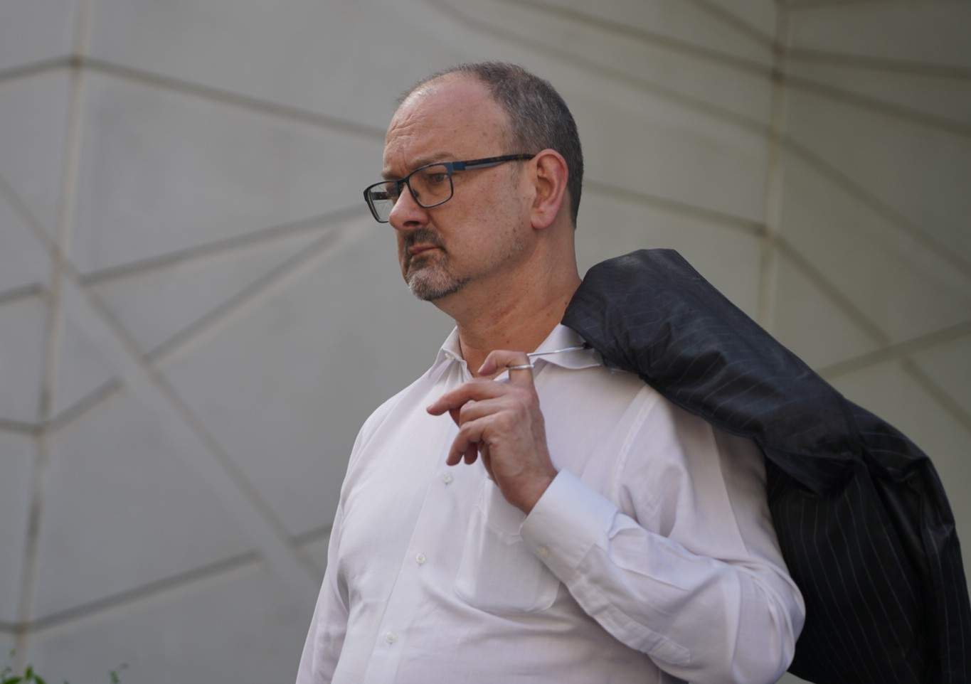A bespectacled man with short hair holds a suit jacket over his shoulder.