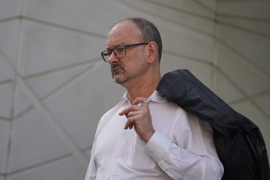 A bespectacled man with short hair holds a suit jacket over his shoulder.