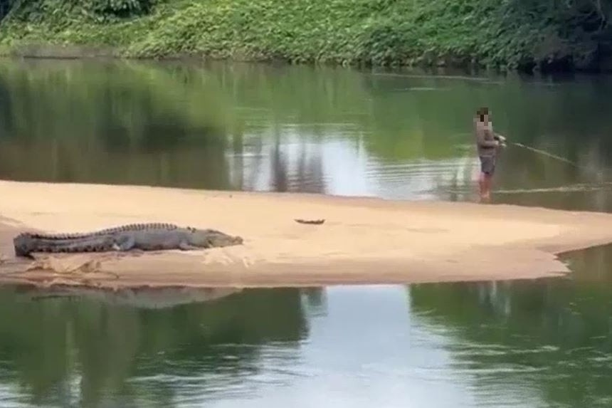 A crocodile sits on a sandy bank while a man fishes nearby