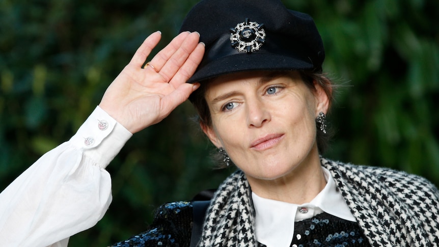 Stella Tennant wearing a black cap holds her hand to her head while looking to the left of camera