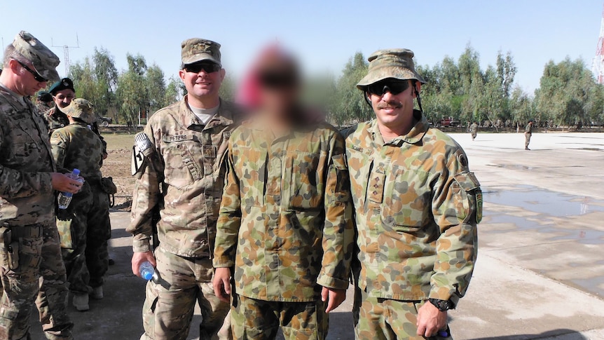 Three men in army fatigues standing together, the middle has his face blurred completely