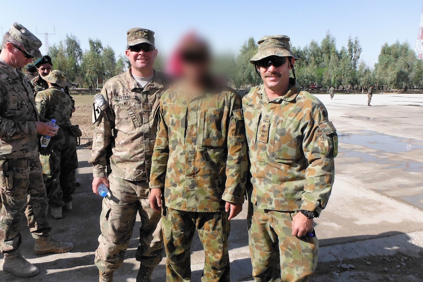 Three men in army fatigues standing together, the middle has his face blurred completely