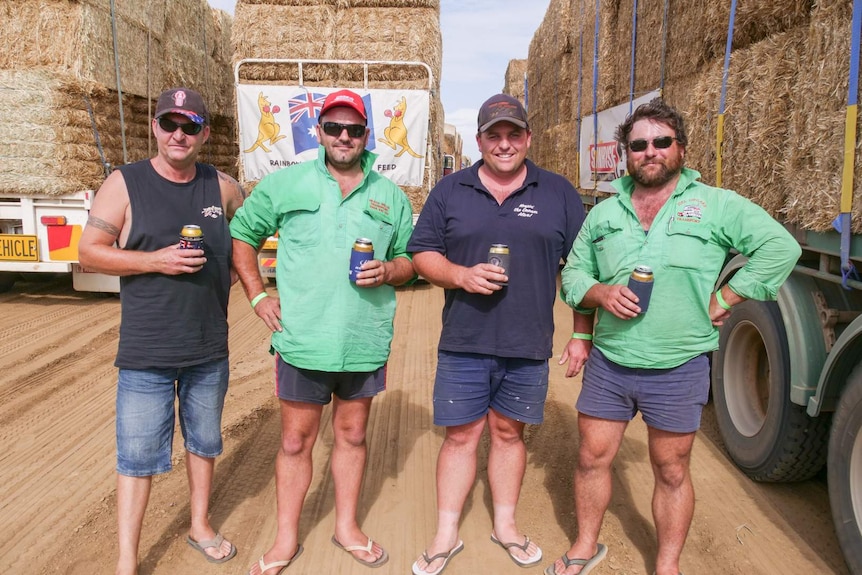 Four men stand in front of many truck of hay, each holding a drink.