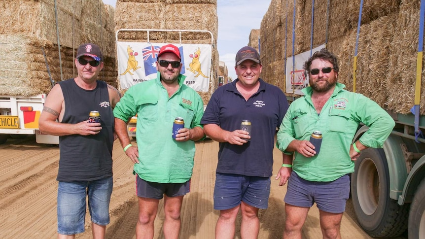 Four men stand in front of many truck of hay, each holding a drink.