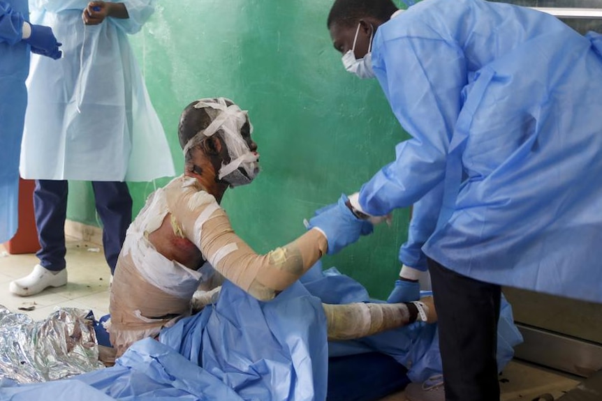 A burns victim sits on the floor wrapped in gauze and bandages as a doctor in blue surgical gown tends to his injuries 