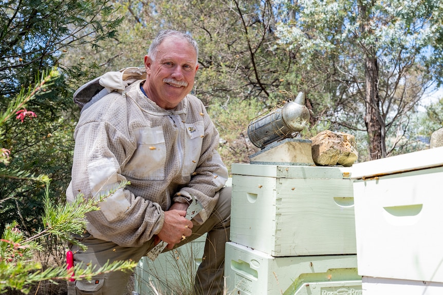Man wearing a beekeeping top kneeling beside a bee hive. 